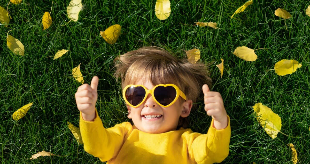 Un enfant allongé dans l'herbe portant des lunettes en cœur souriant à la caméra sous une pluie de feuilles en levant les deux pouces